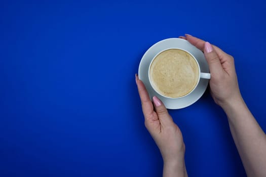 Women's hands hold a white mug with hot coffee. Blue background. Space for text. Mockup. Blue background. A place to copy.