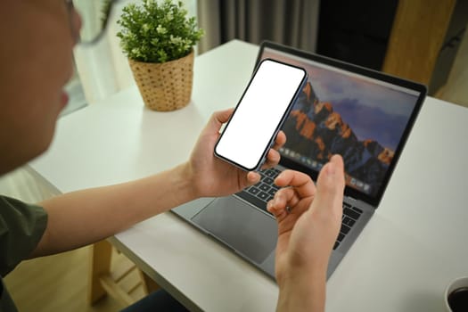 Closeup view of man sitting in front of laptop at working desk and using smartphone.