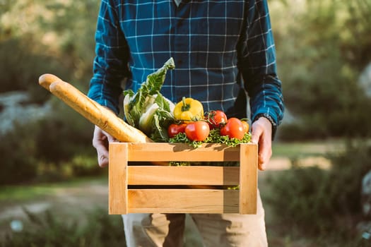 Caucasian man male holding wooden crate with vegan groceries organic vegetables and bread baguette. Farmer delivered a framed box with supplies to a farmers market camping site in the forest. No face.