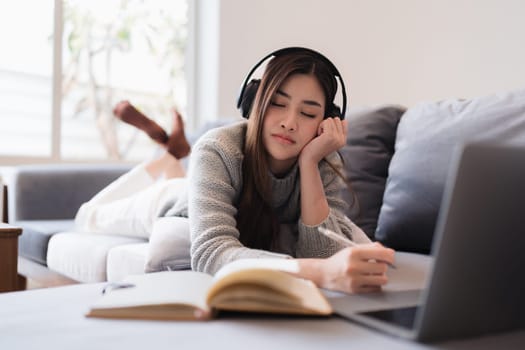 Online education, e-learning. Asian woman in stylish casual clothes, studying using a laptop, listening to online lecture, taking notes, online study at home.