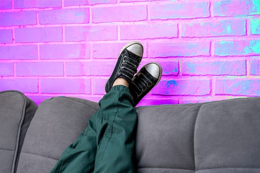 Relaxed legs of a boy in green casual pants and comfortable sneakers on a cozy sofa in the living room against a neon violet brick wall. children's feet in fashionable sneakers lie on the back of the sofa. Space for copying. Light violet neon brick background