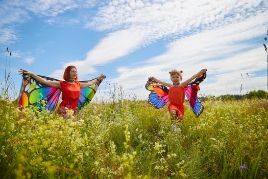 Happy female family with red haired mother and daughter with bright butterfly wings having fun on green and yellow meadow full of grass and flowers in sunny summer day. Concept family love