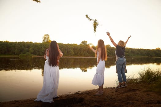 Three female girls in white sundresses and with flower wreaths on the riverbank at sunset. The feast of Ivan Kupala and national clothes and traditions of throwing wreath into the water