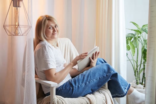 Young blonde woman with book on chair near window at home. Student girl is resting and reading textbook at home. Nice atmosphere. The concept of cozy home