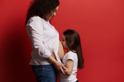 Happy multi ethnic gravid woman standing bare belly with her little daughter kissing her tummy, isolated on red background. Mom and daughter together. Expecting baby. Family relationships and values