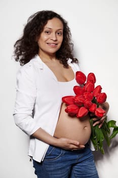 Multi ethnic pregnant woman posing bare belly with a bouquet of red tulips, touching her tummy, expressing positive emotions from pregnancy and maternity, smiling at camera, isolated white background