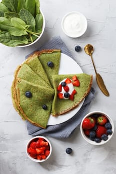 Spinach green pancakes on a plate with fresh berries and sour cream, on a gray background with textiles. vertical photo