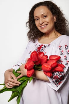 Beautiful woman in embroidered white dress, holds a bouquet of red tulips, smiles looking at camera, isolated on white background. Copy space. Ukrainian style, culture and traditions. Independence Day