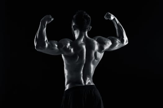 Image of very muscular man posing with naked torso in studio on black background. Rear view. Black and white color