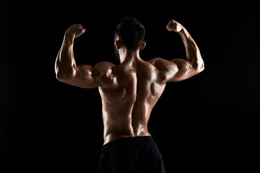 Image of very muscular man posing with naked torso in studio on black background. Rear view