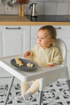 Happy baby sitting in high chair eating carrot in kitchen. Healthy nutrition for kids. Bio carrot as first solid food for infant. Children eat vegetables. Little boy biting raw vegetable