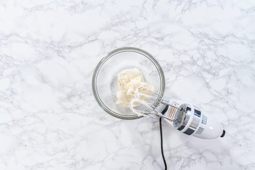Flat lay. Mixing store-bought vanilla frosting in a mixing glass bowl with a hand mixer.