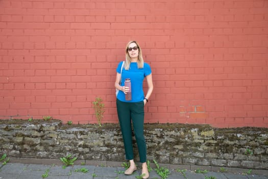 a blonde in a blue blouse against a brick wall drinks water from sports water bottle with time markers, to comply with the drinking regime, High quality photo