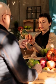 Young woman farmer standing behind farmers market stall showing organic locally grown carrots to customer while selling fresh fruits and vegetables. Mature man buying healthy local food