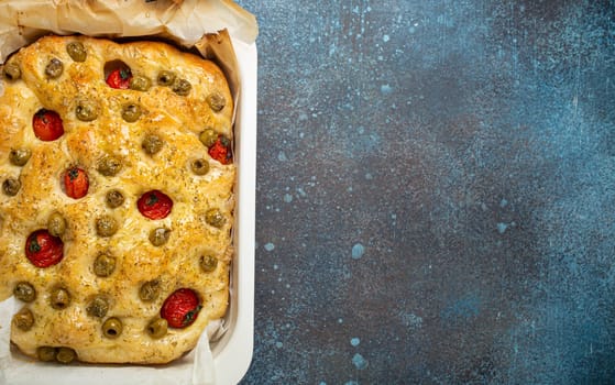 Overhead of Traditional Italian Homemade Flat Bread Focaccia with Green Olives, Olive Oil, Cherry Tomatoes and Rosemary in Baking Tray on Rustic Dark Blue Stone Background, Space for Text