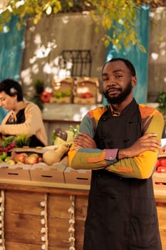Successful eco-friendly business. Young confident African American guy farmer wearing apron looking at camera while standing at local market and waiting for clients, selling fresh organic produce