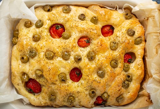 Overhead of Traditional Italian Homemade Flat Bread Focaccia with Green Olives, Olive Oil, Cherry Tomatoes and Rosemary in Baking Tray on Rustic Dark Blue Concrete Background