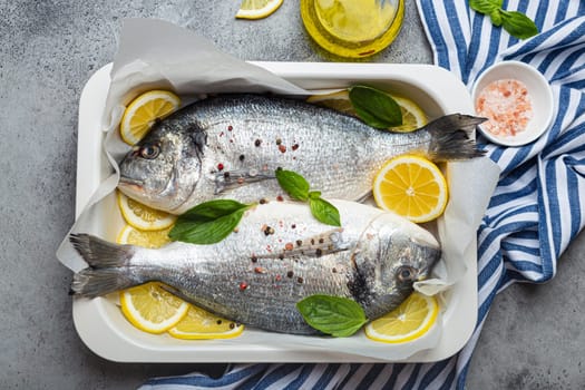 Raw fish dorado in casserole dish with ingredients lemon, fresh basil, bottle of olive oil on wooden cutting board with knife on rustic stone background top view, cooking healthy fish dorado concept