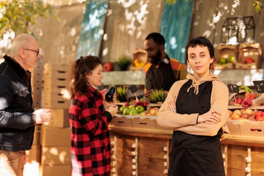 Young confident woman wearing apron running successful farmers market stand, selling fresh organic produce directly to customers. Female greengrocer standing with crossed arms and looking at camera