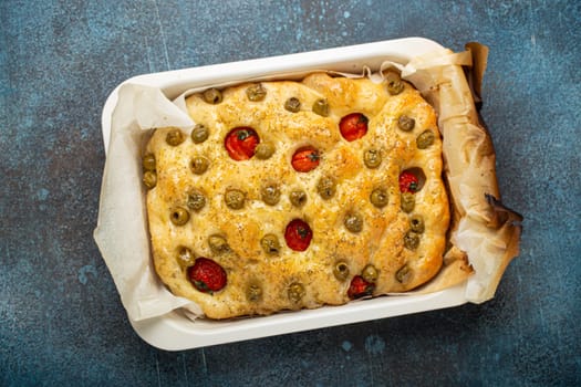 Overhead of Traditional Italian Homemade Flat Bread Focaccia with Green Olives, Olive Oil, Cherry Tomatoes and Rosemary in Baking Tray on Rustic Dark Blue Concrete Background