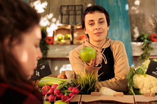 Young pleasant female farmer standing behind market stand with healthy organic produce holding green apple and smiling at camera. Woman selling fresh locally grown fruits and vegetables