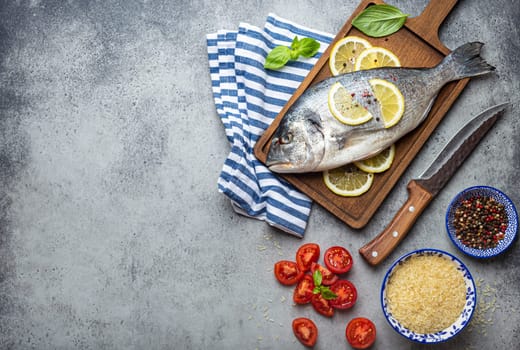 Raw fish dorado with ingredients lemon, fresh basil, cut cherry tomatoes, uncooked rice on wooden cutting board with knife on rustic stone background top view, cooking healthy fish dorado. Copy space