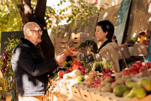 Woman seller showing organic natural carrots to elderly man at farmers market, locally grown. Small business owner standing behind fruit and vegetable stall selling healthy fresh produce to customer.