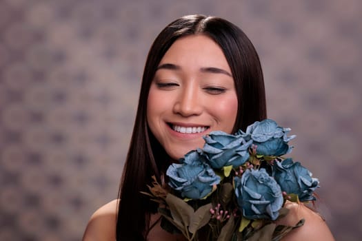 Grateful happy smiling asian woman holding blue roses bouquet and looking at flowers blossom. Young beauty industry model posing with bunch of blooming plants in hands close up