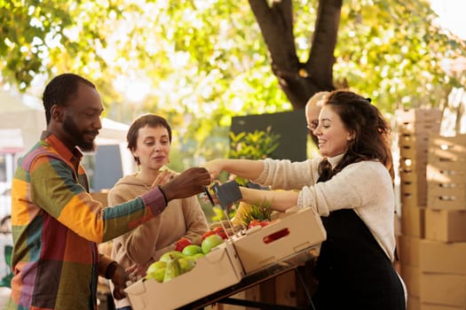 Vendor offering samples to customers while selling home-grown fruits and vegetables at local farmers market. Young multiracial family couple tasting natural organic produce while visiting food fair