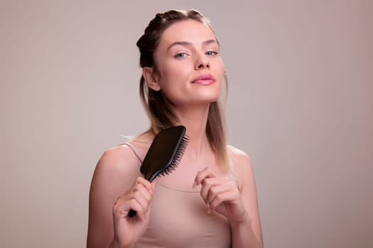 Woman brushing healthy blonde hair and looking at camera. Attractive young fashion model wearing natural make up and casual beige top using hairbrush for hairstyling routine
