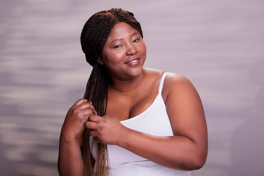 Natural beautiful african american woman making braids hairstyle, wearing white top, smiling and looking at camera. Attractive body positive model showing haircare daily procedure