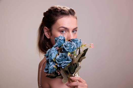 Young woman holding blue roses bouquet in front of face and looking at camera. Fashion model posing with unique flowers bunch in hand for creative portrait on beige background