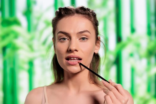 Young model doing lips make up with brush portrait. Attractive woman with healthy fresh skin applying lipbalm, showing makeup daily routine and looking at camera on bamboo background