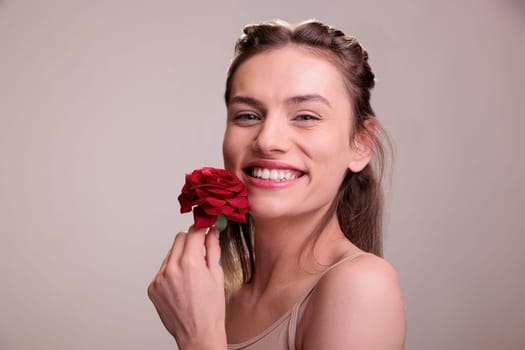 Laughing young woman posing with red rose gift portrait. Attractive cheerful caucasian blonde lady wearing nude makeup holding flower romantic present and expressing positive emotion