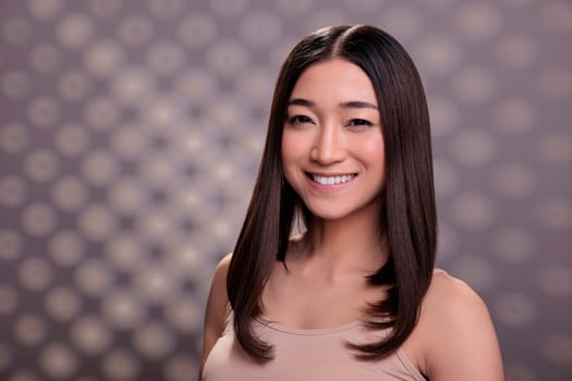 Pretty asian optimistic woman smiling and looking at camera face portrait. Beauty model with healthy fresh skin wearing beige casual top and natural cosmetics make up posing in studio
