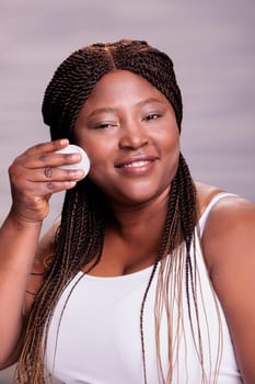 Plus size smiling model using cotton pad disk for facial makeup cleaning portrait. Body positive african american woman doing daily skincare routine cleansing step, washing face