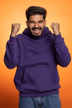 Happy joyful indian man shouting, celebrating success winning, birthday, lottery jackpot goal achievemen, good news, victory, watching TV championship. Young hindu guy on orange studio wall background