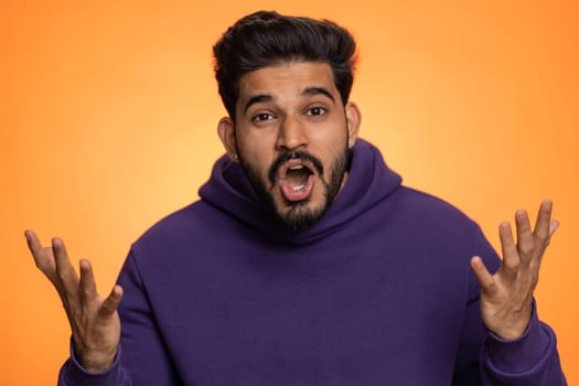 Oh my God, Wow. Excited amazed winner man raising hands in surprise looking at camera with big eyes, shocked by sudden victory. Sincere handsome indian guy isolated alone on orange studio background