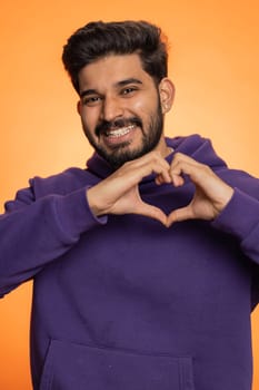 Man in love. Smiling bearded indian man 25 years old makes heart gesture demonstrates love sign expresses good feelings and sympathy. Handsome hindu young guy isolated on orange studio background