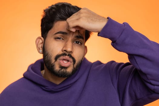 Handsome indian bearded man in hoodie rubbing temples to cure headache problem, suffering from tension, migaine, stress, grimacing in pain, high blood pressure isolated alone on orange wall background