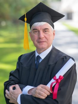 Portrait of an elderly man in a graduation gown and with a diploma in his hands outdoors. Vertical