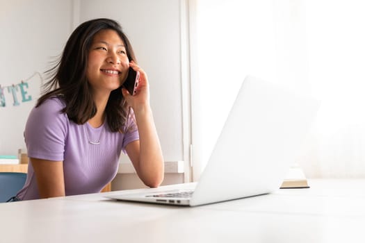 Happy Asian woman smiling talking on the phone while working at home office. College student using mobile phone. Technology concept.