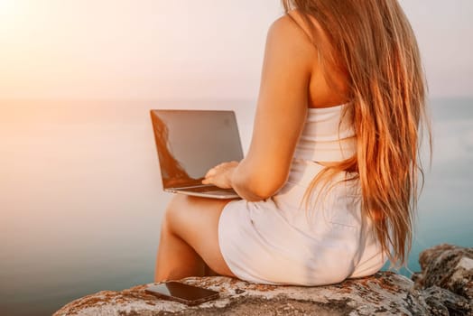 Digital nomad, Business woman working on laptop by the sea. Pretty lady typing on computer by the sea at sunset, makes a business transaction online from a distance. Freelance, remote work on vacation