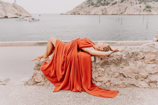 Side view a Young beautiful sensual woman in a red long dress posing on a volcanic rock high above the sea during sunset. Girl on the nature on overcast sky background. Fashion photo