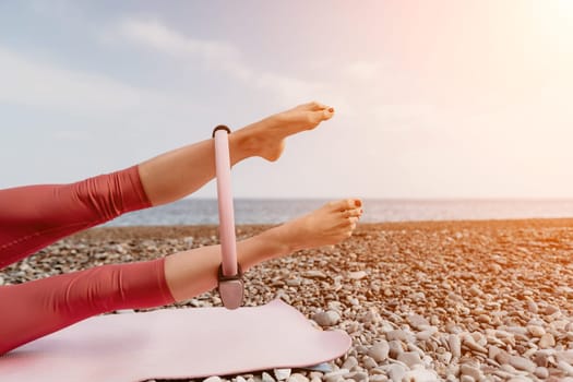 Middle aged well looking woman with black hair doing Pilates with the ring on the yoga mat near the sea on the pebble beach. Female fitness yoga concept. Healthy lifestyle, harmony and meditation.