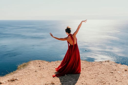 Side view a Young beautiful sensual woman in a red long dress posing on a rock high above the sea during sunrise. Girl on the nature on blue sky background. Fashion photo.