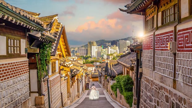 Girl wearing hanbok walking through Bukchon Hanok Village with Seoul city skyline, cityscape of South Korea at sunset