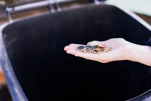 A kid girl hand throws euro coins into a trash can dumpster, street shot.