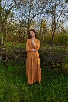 an elegant, sophisticated woman poses relaxed standing near a wicker fence at the dacha in a long orange dress enjoying the silence and peace. High quality photo