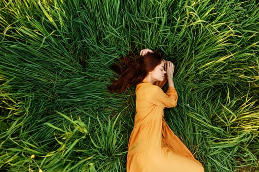 an elegant woman lies in the tall green grass in a long summer orange dress and holding her hair with her hands enjoys nature and sunny weather with her eyes closed. High quality photo